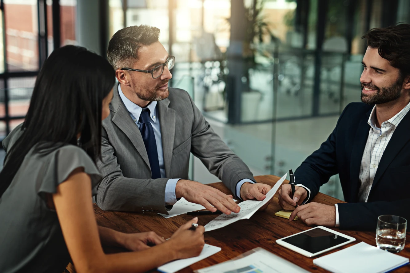 Three business people discussing documents around a wooden table in an office.